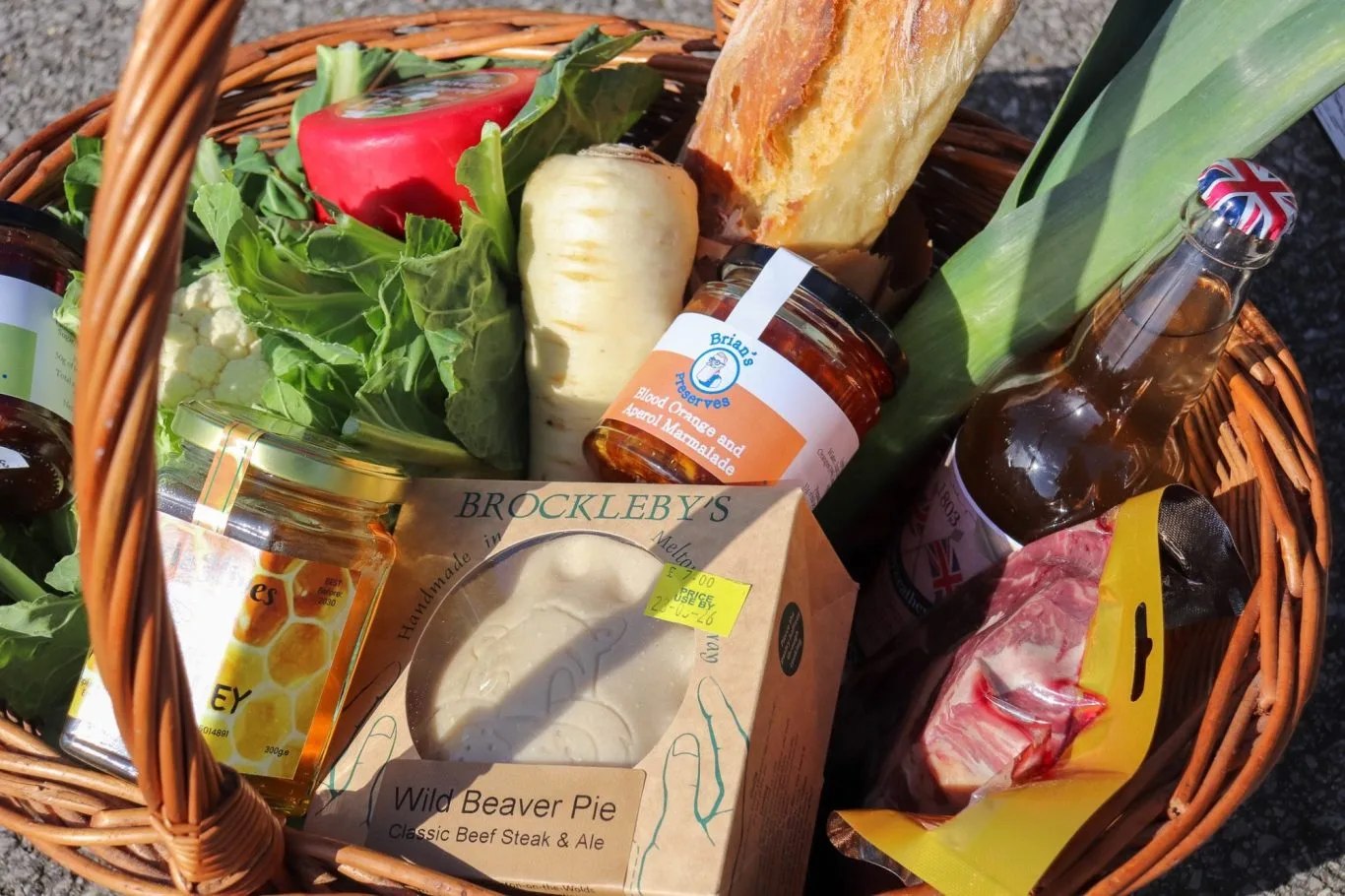 A wicker basket filled with local produce: vegetables, preserves, a pie, cheese and bottled drinks