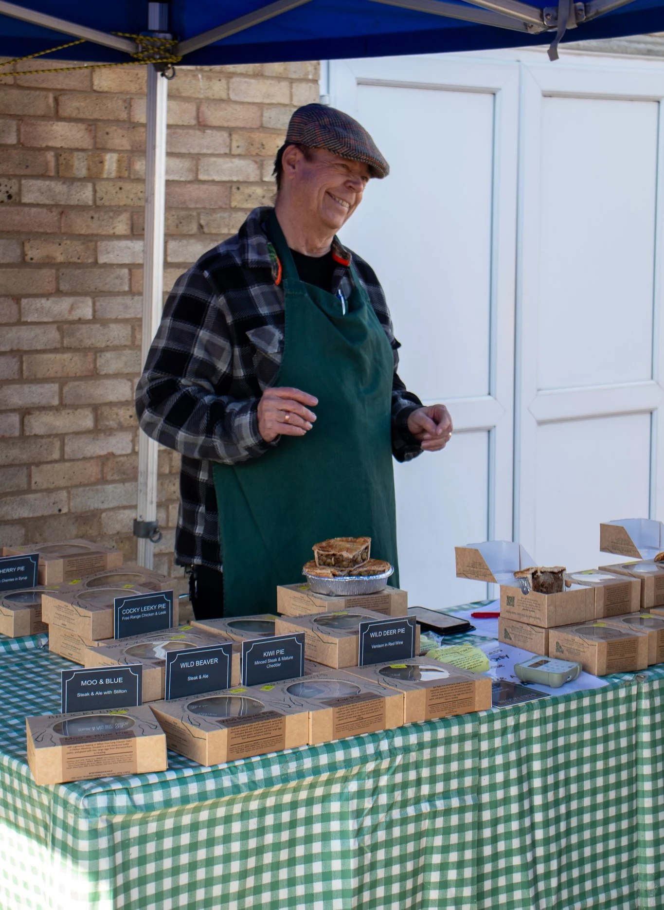 A smiling pie trader in a green apron behind his stall of boxed pies